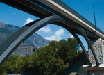 Photo: view from below on bridge; behind trees, buildings, mountains; blue sky with scattered clouds
